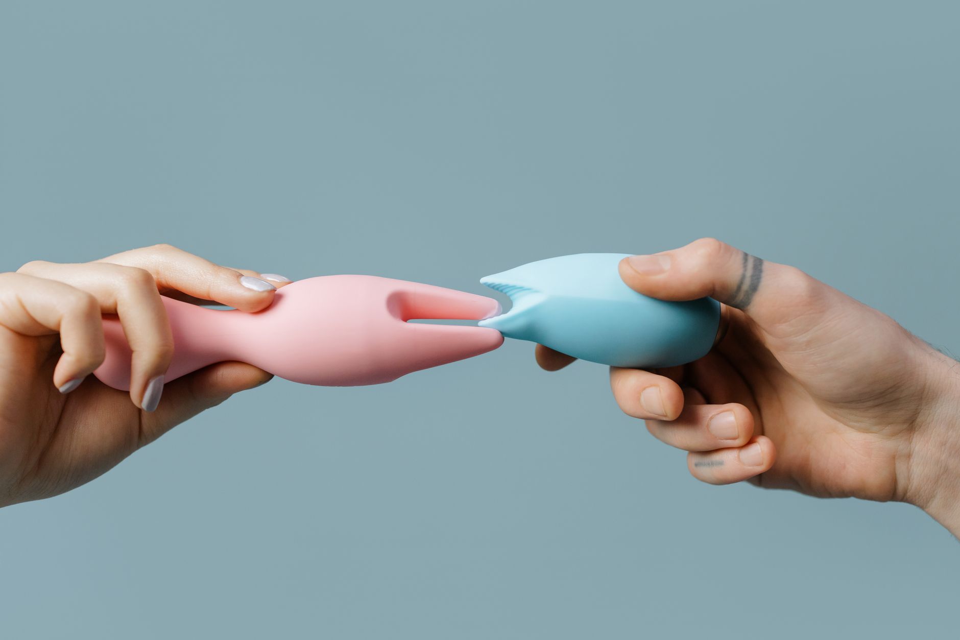 Two hands holding pink and blue silicone vibrators against a soft pastel background